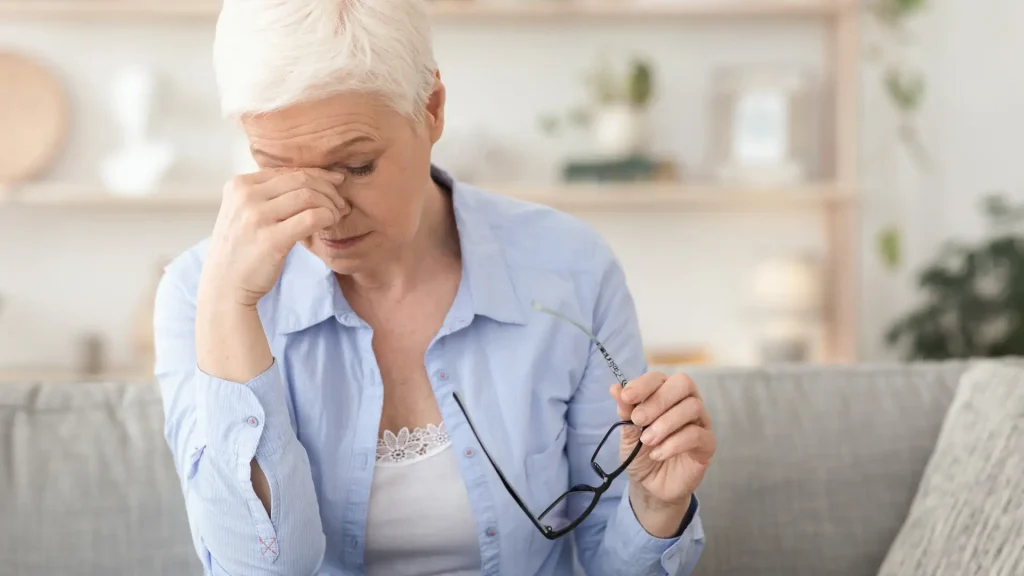 Older woman holding glasses and pinching the bridge of her nose, appearing fatigued.