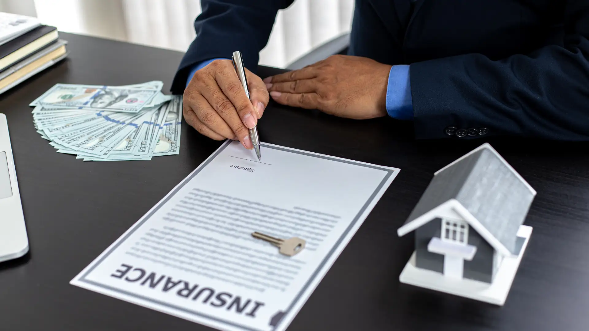 Person signing an insurance document with money, keys, and a small house model on the desk.